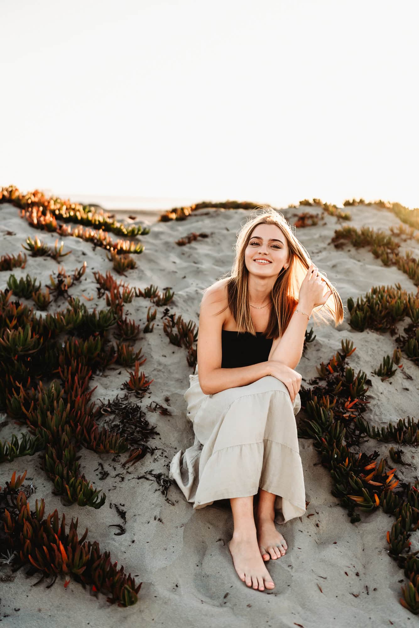 A young blond woman sits on a sand dune on Coronado Beach during her senior portraits session, playing with her hair and smiling.