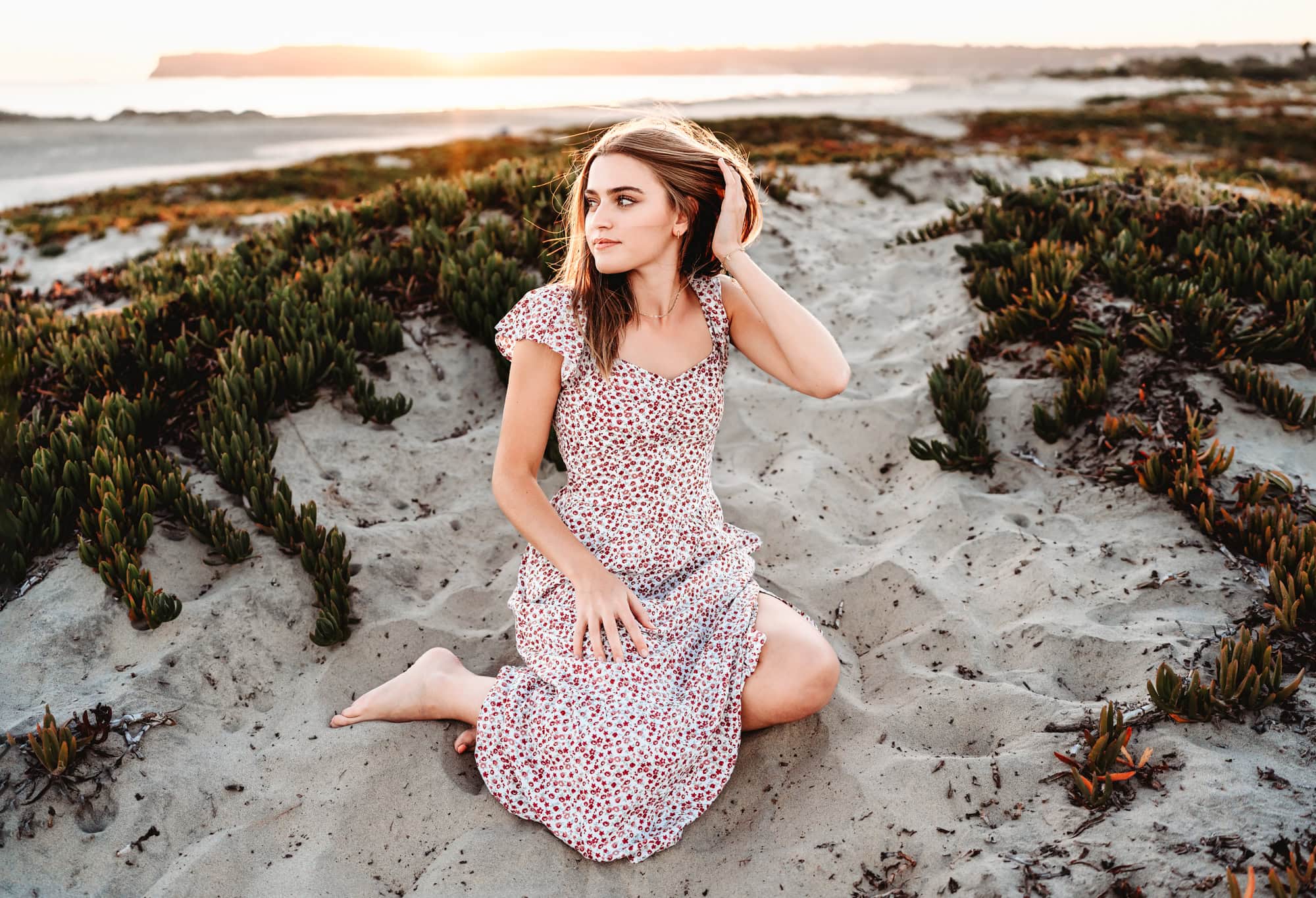 A senior girl sits on a San Diego sand dune during her senior photoshoot on Coronado Beach.