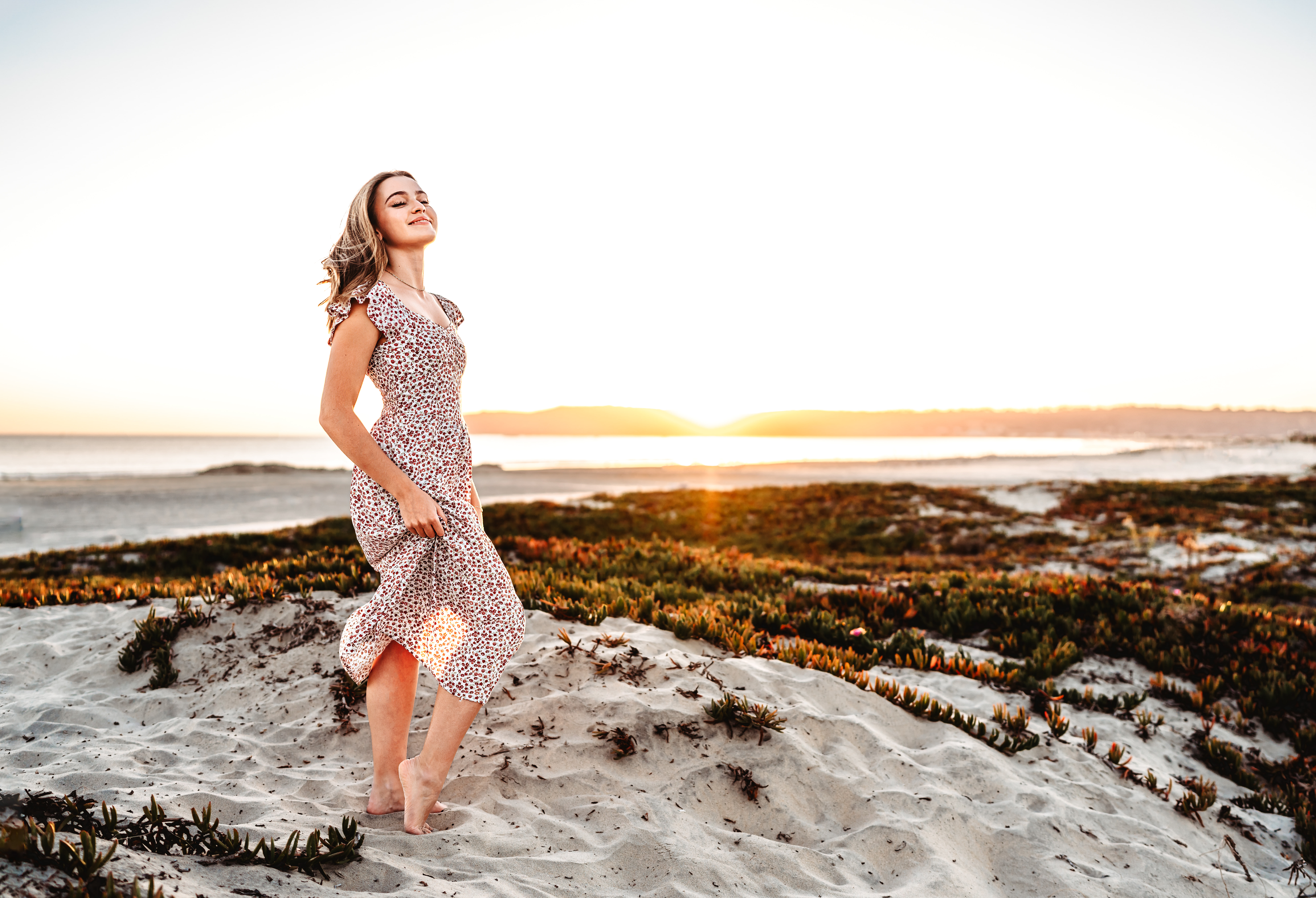 A senior girl stands on a Coronado Beach sand dune, holding her dress and flipping her hair, during a San Diego beach sunset senior session. 