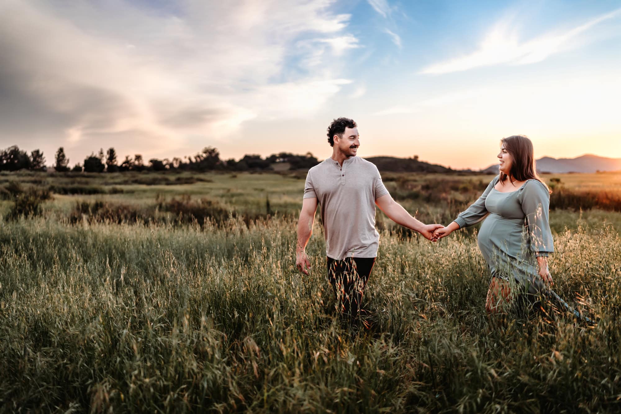 Golden hour maternity photo in San Diego featuring an expectant couple walking hand-in-hand through tall grass, bathed in warm sunset light.