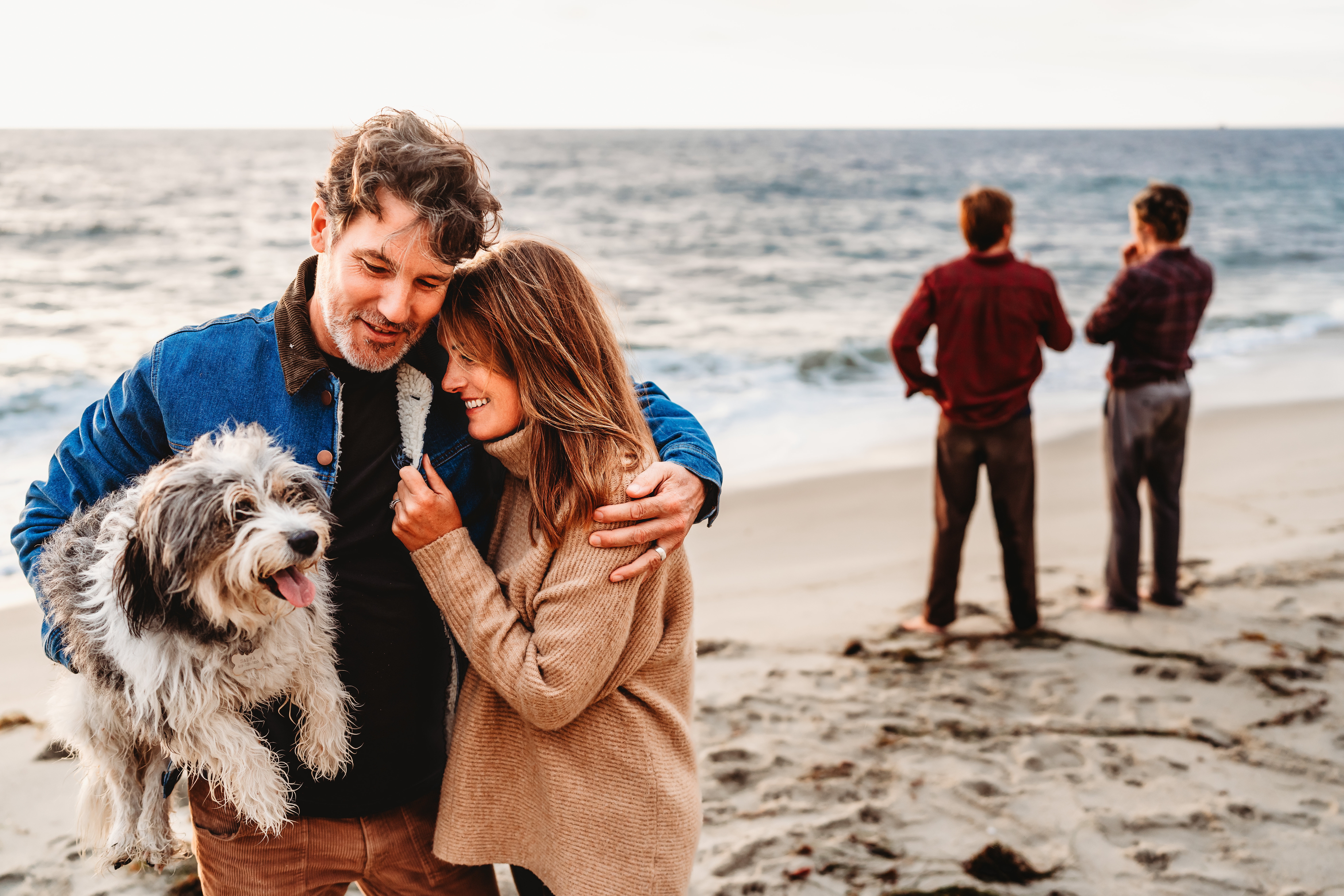 A man holding his white dog on a La Jolla beach embraces his wife in a picture illustrating what to wear for fall family photos in San Diego. 
