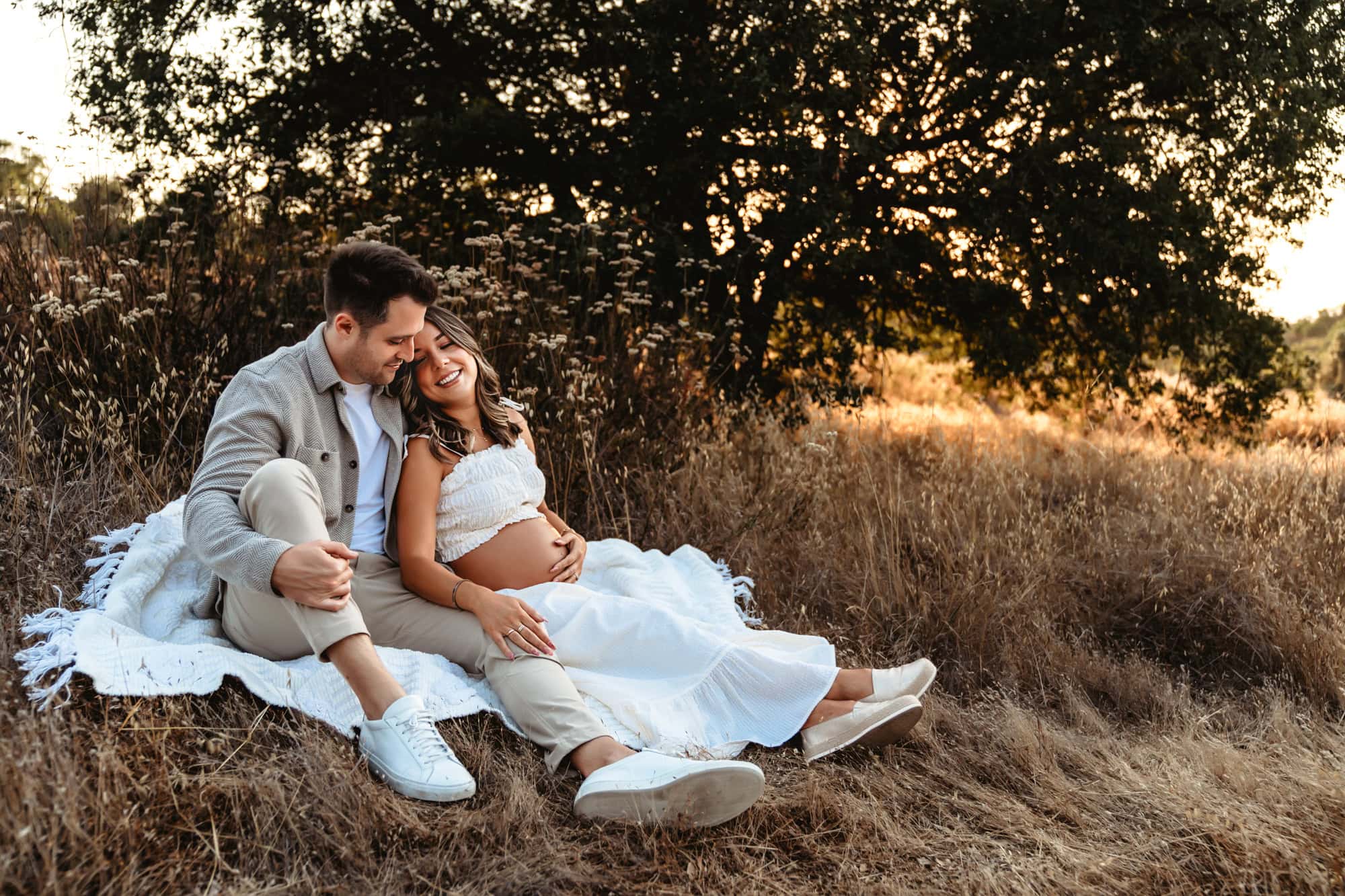 A man and his pregnant wife nestle together on a blanket next to a tree during their maternity photoshoot in San Diego.