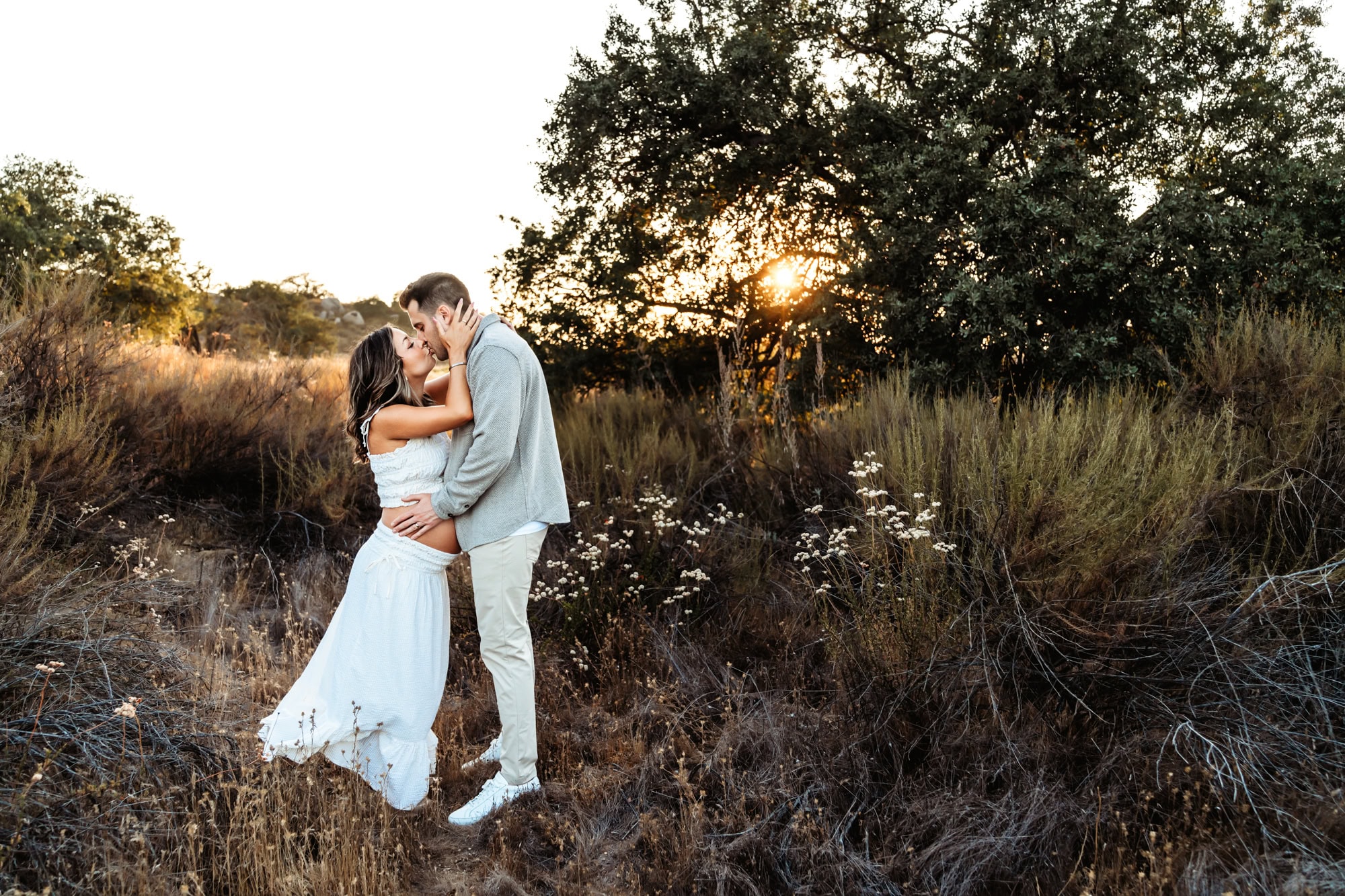 A man and his pregnant wife kiss each other in a San Diego field at sunset.