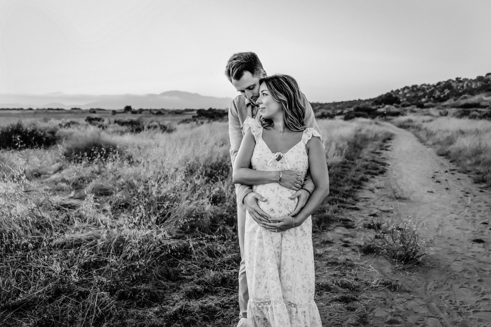 A man hugs his pregnant wife from behind as she looks off toward the sunset while having maternity photos taken in San Diego. 