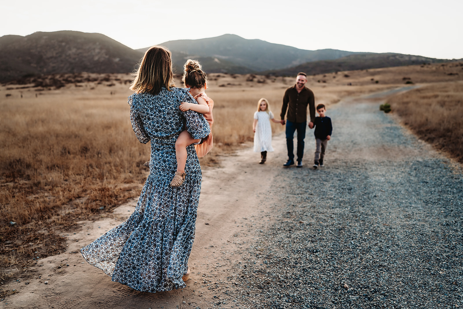 A woman hold her toddler daughter while watching her husband and kids walk toward her in Mission Trails Regional Park, in an article about what to wear for fall family photos in San Diego. 