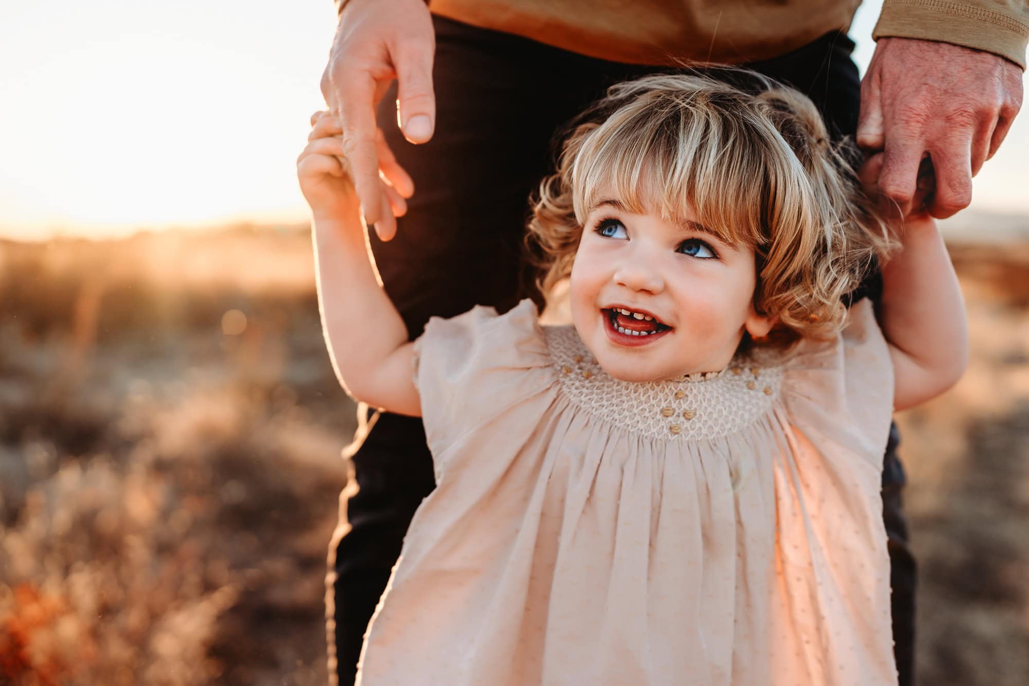 A toddler girl holds her dad's hands and looks off, laughing, during a sunset family photo session in a San Diego field. 