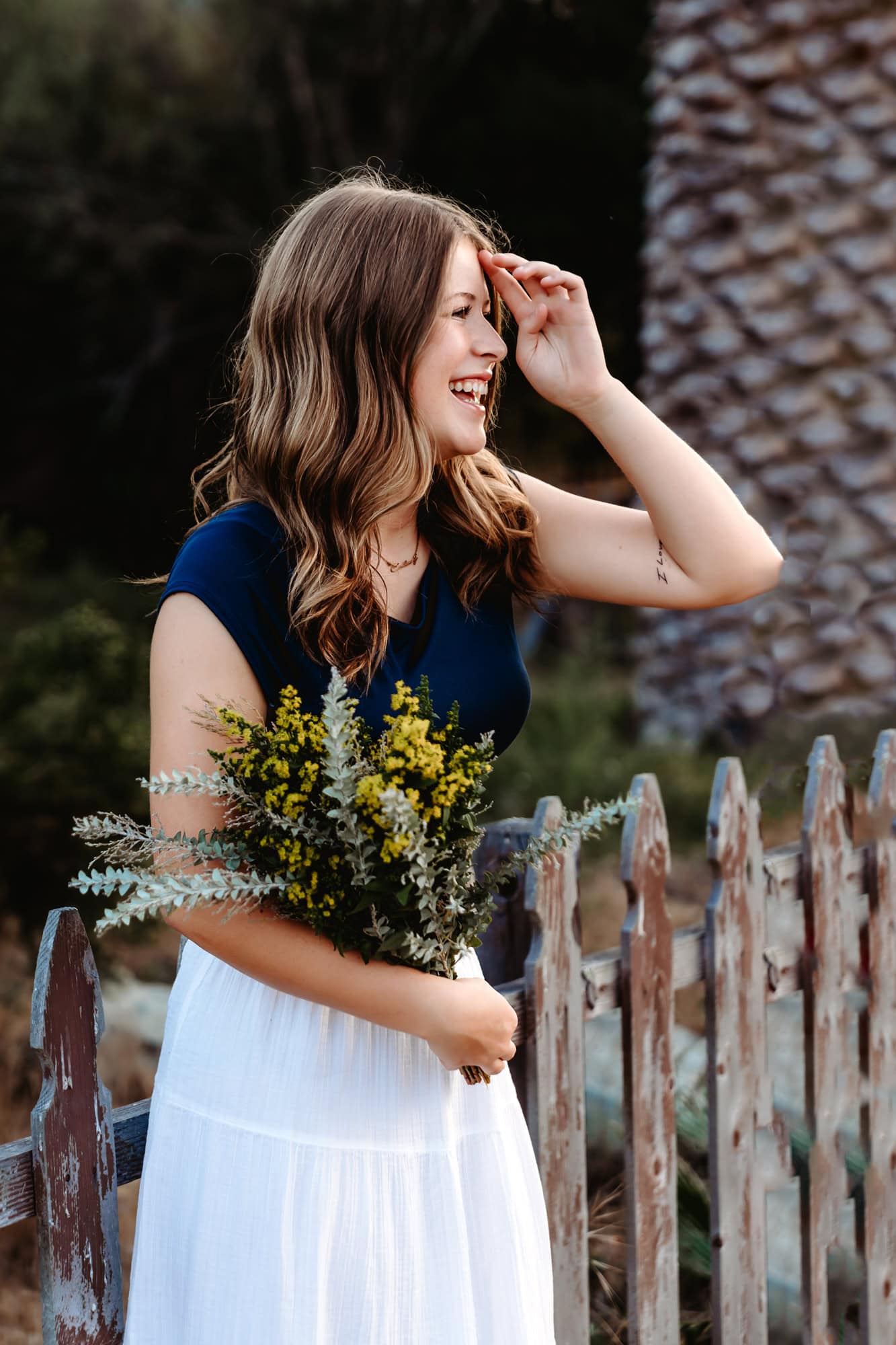 High school senior standing by a weathered wooden fence holding flowers in natural outdoor senior pictures in San Diego.