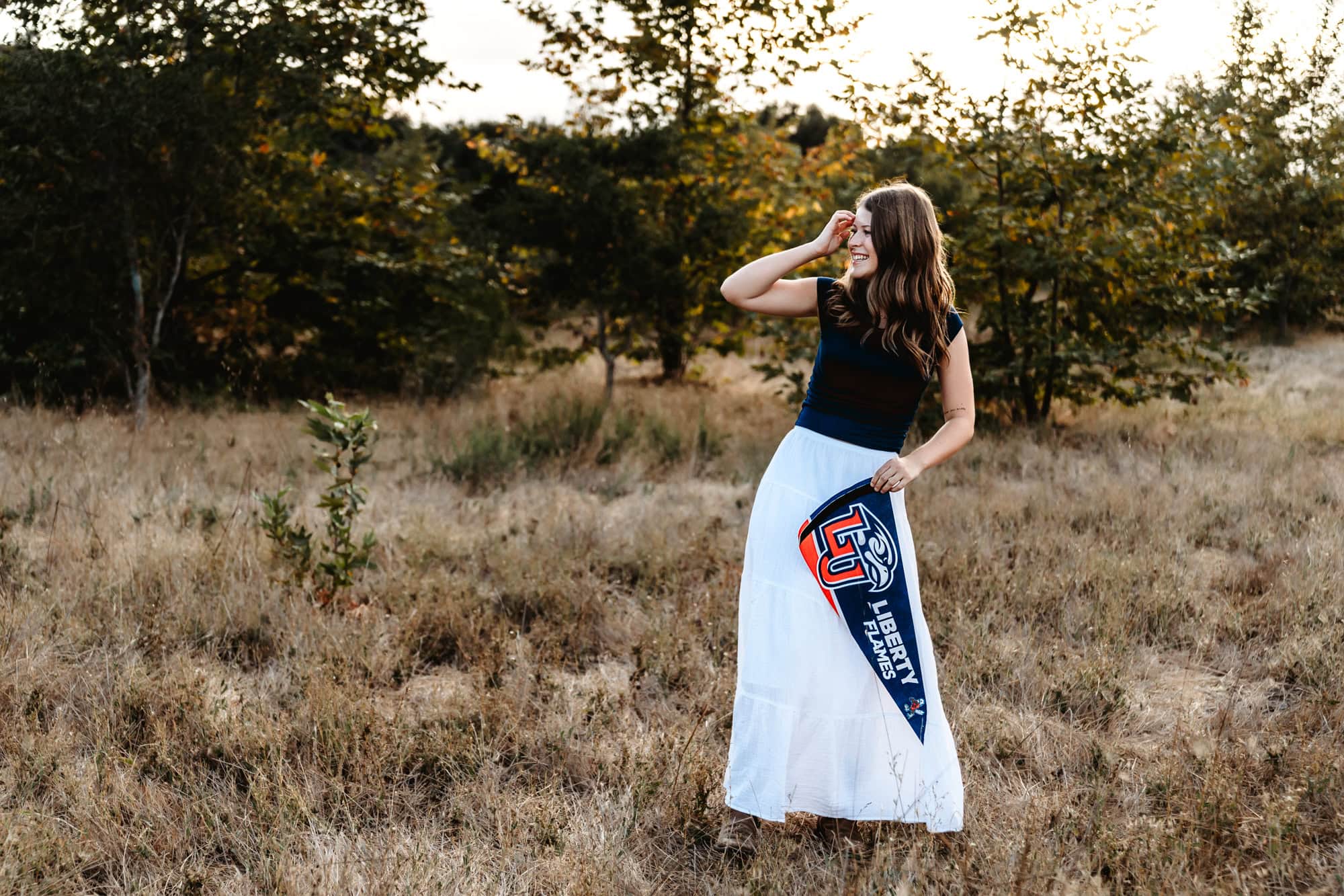 Senior girl standing in an open grassy field, holding her college pennant while wearing a long skirt for golden hour senior portraits in San Diego.