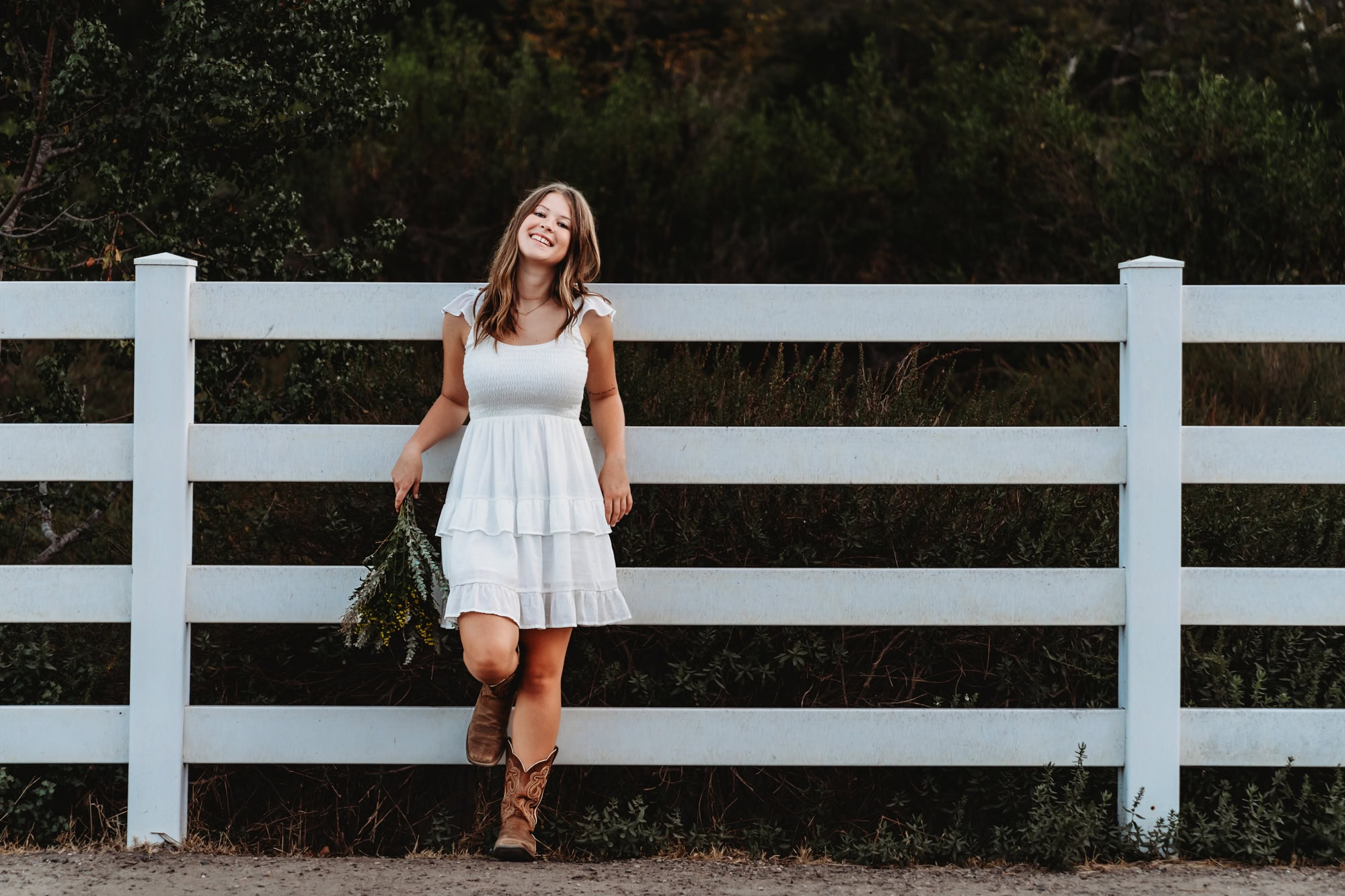 Senior girl in a cream dress leaning against white fence, rustic and natural senior portraits in San Diego.