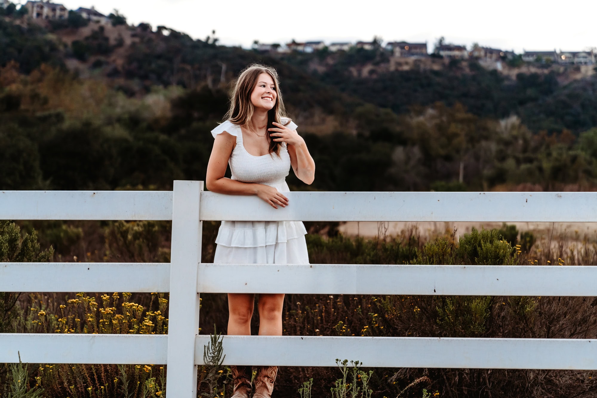 Senior portrait of girl in a cream dress climbing a fence at Los Peñasquitos Gardens sign, playful San Diego senior pictures.