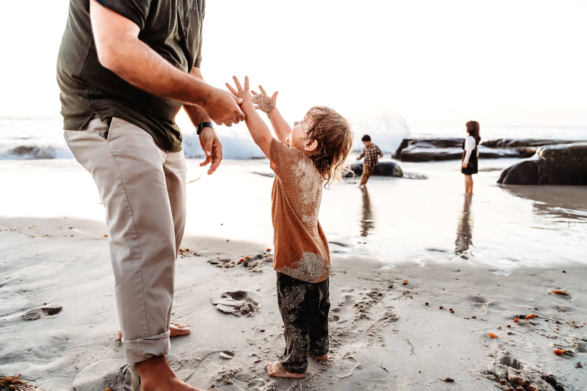 Father and young son playing together near the water’s edge during a family photoshoot on Windansea Beach. The boy is lifting his hands up, begging his father to pick him up.