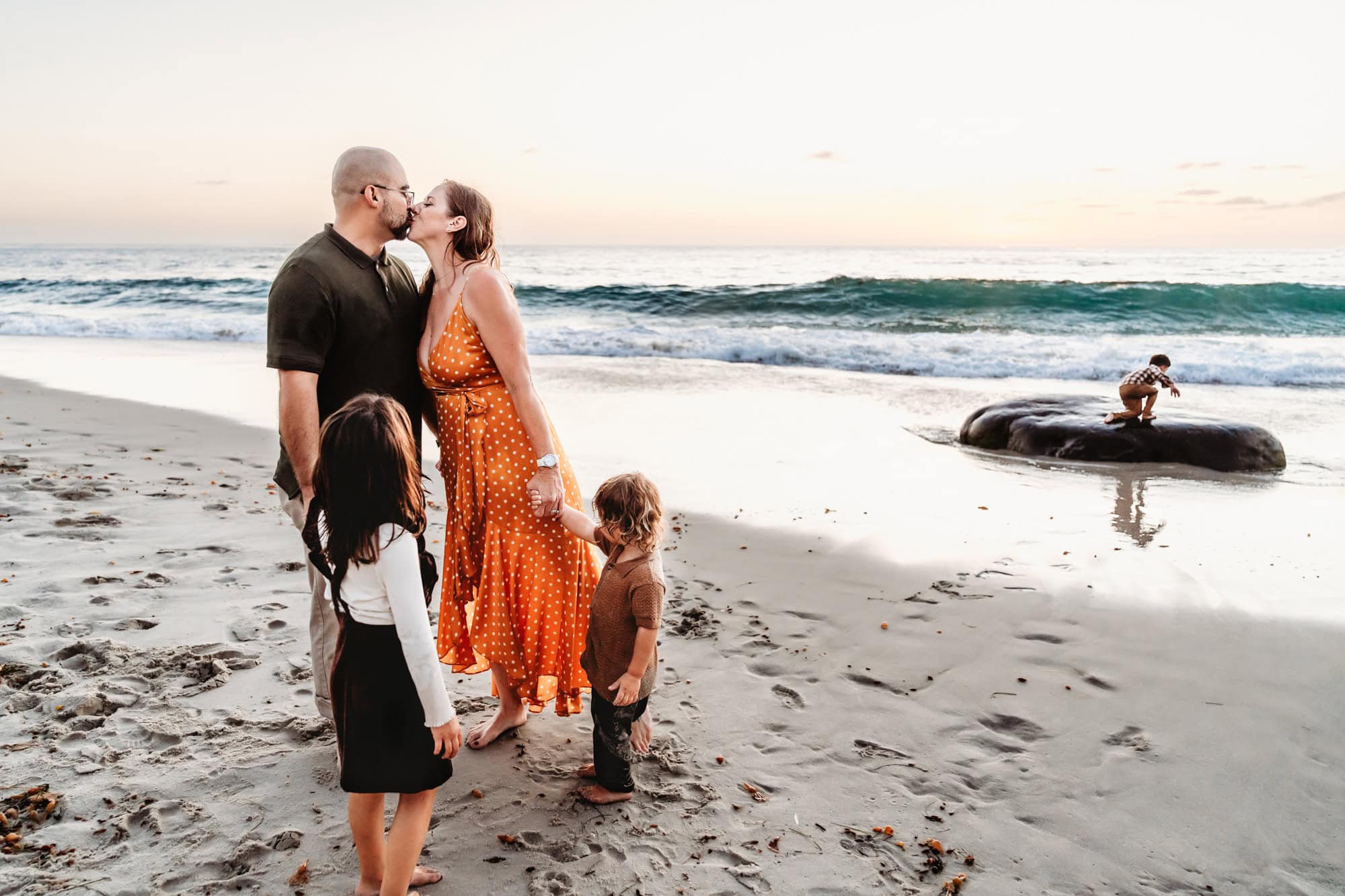 A mom and dad kiss during a beach family photo session in La Jolla, CA while their son plays in the background. 