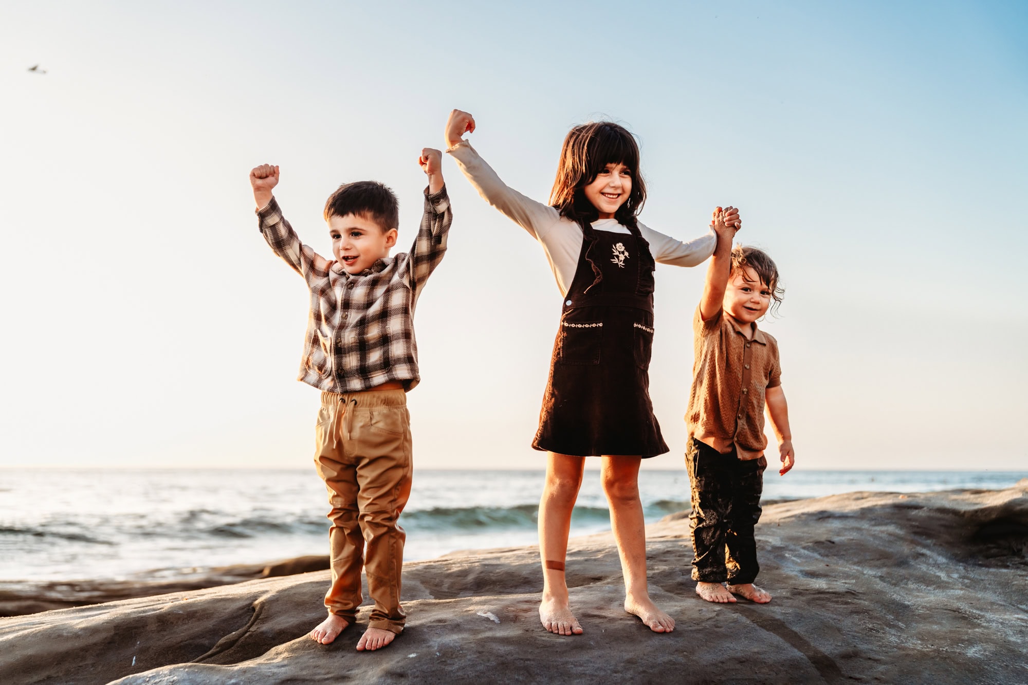 Three young siblings flexing their muscles and laughing together on beach rocks at sunset in San Diego. 