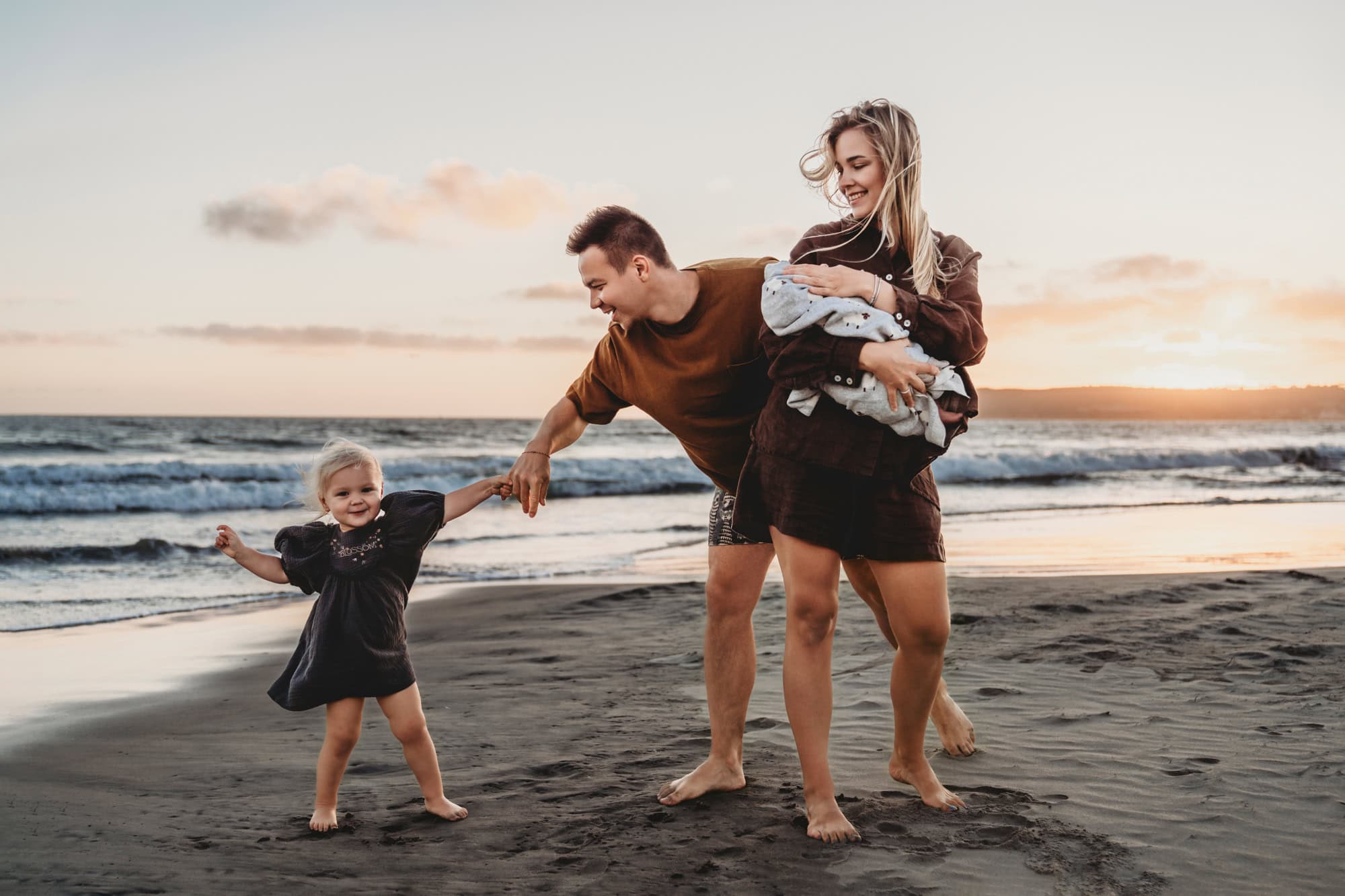 Family playing on a San Diego beach at sunset, with dad playfully guiding his toddler while mom holds a newborn and smiles.