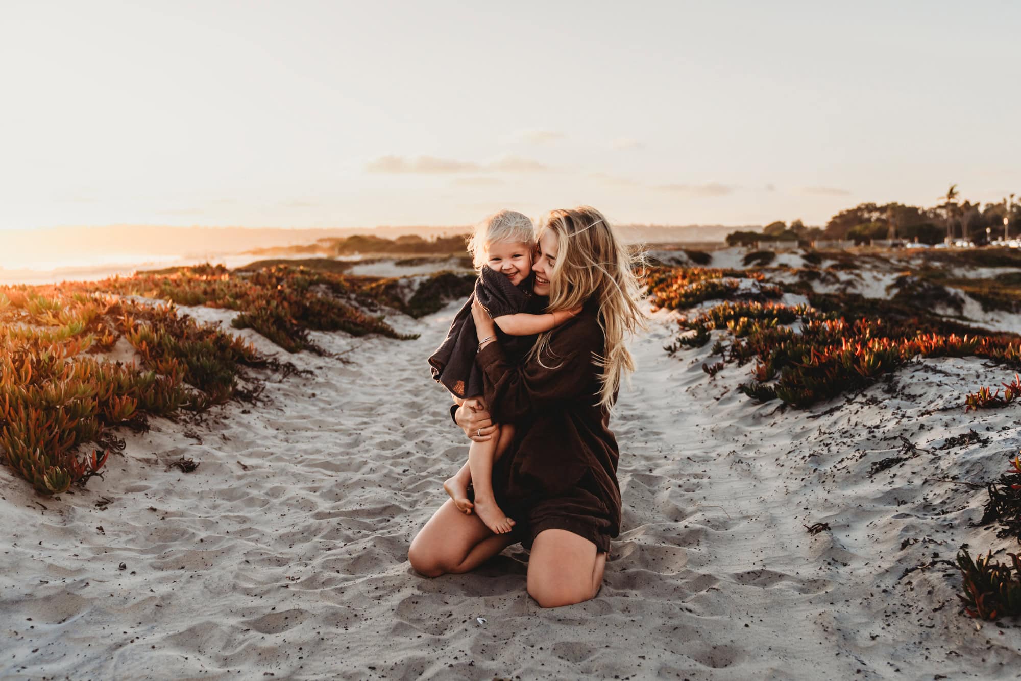 Mother kneeling in the sand at a San Diego beach, hugging her laughing toddler during golden hour with dunes and coastal plants behind them.