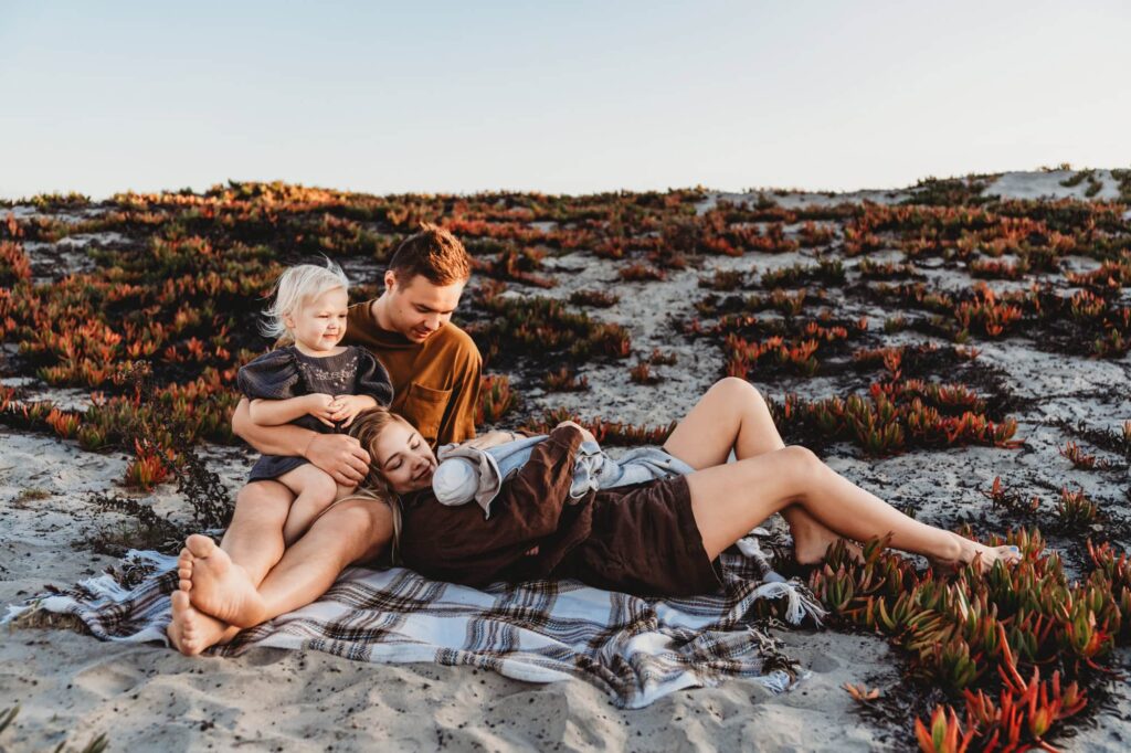 Lifestyle newborn session at Coronado Beach with a San Diego family cuddling their newborn and toddler on a blanket in the sand.