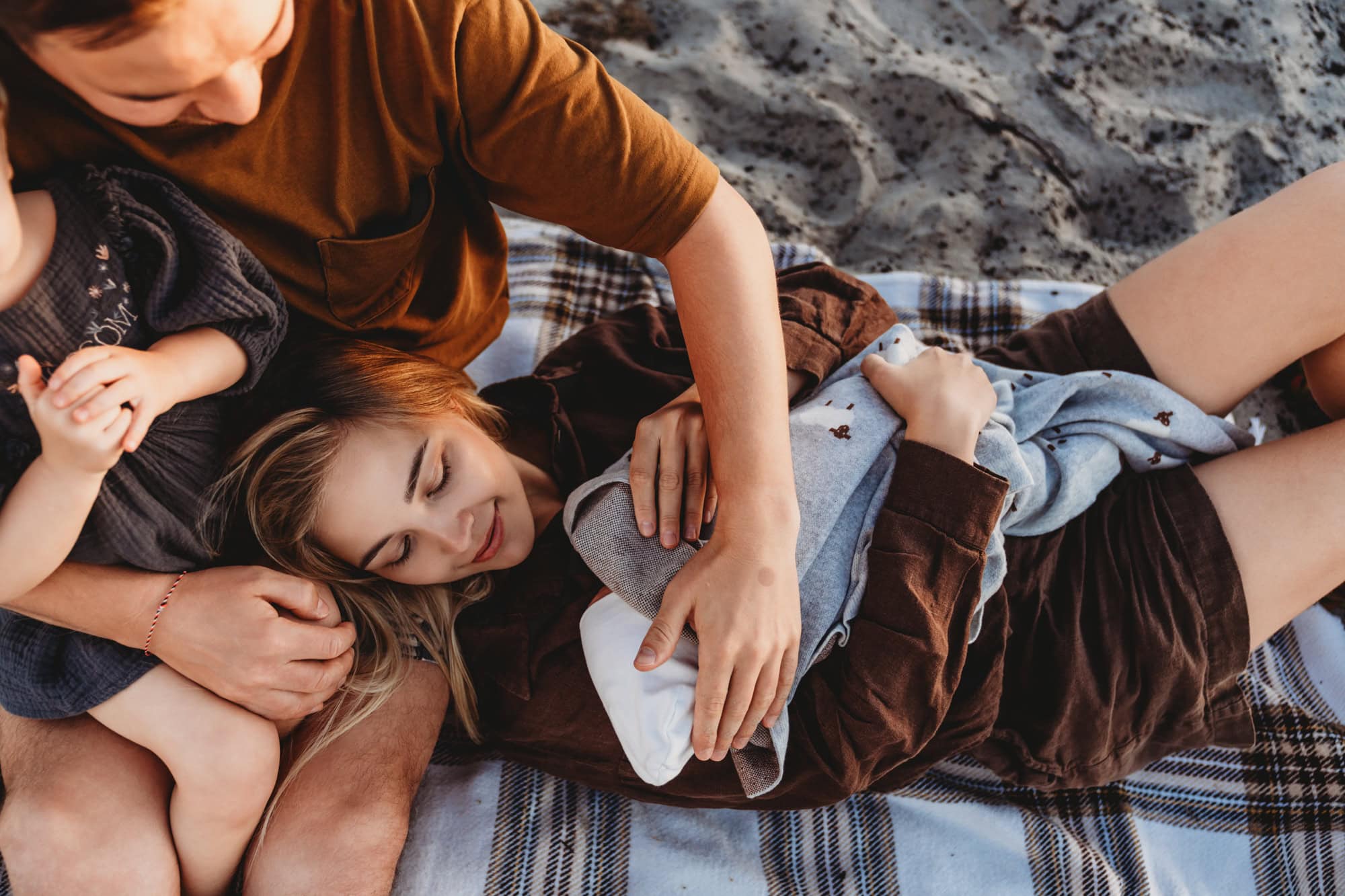 Close-up of parents relaxing on a beach blanket, gently cradling their newborn during an outdoor Coronado Beach session in San Diego.