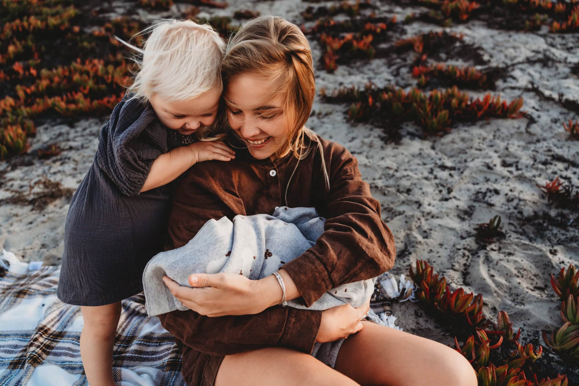 San Diego mom smiling with her toddler leaning in and newborn in her arms, photographed on the dunes at Coronado Beach at sunset.