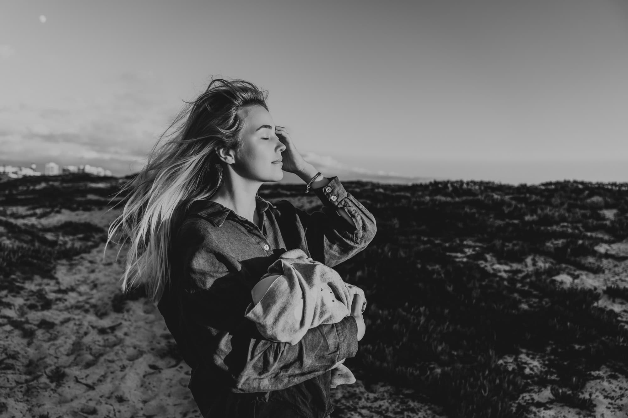 Black and white portrait of a mother holding her newborn against the coastline at Coronado Beach by a San Diego lifestyle newborn photographer.