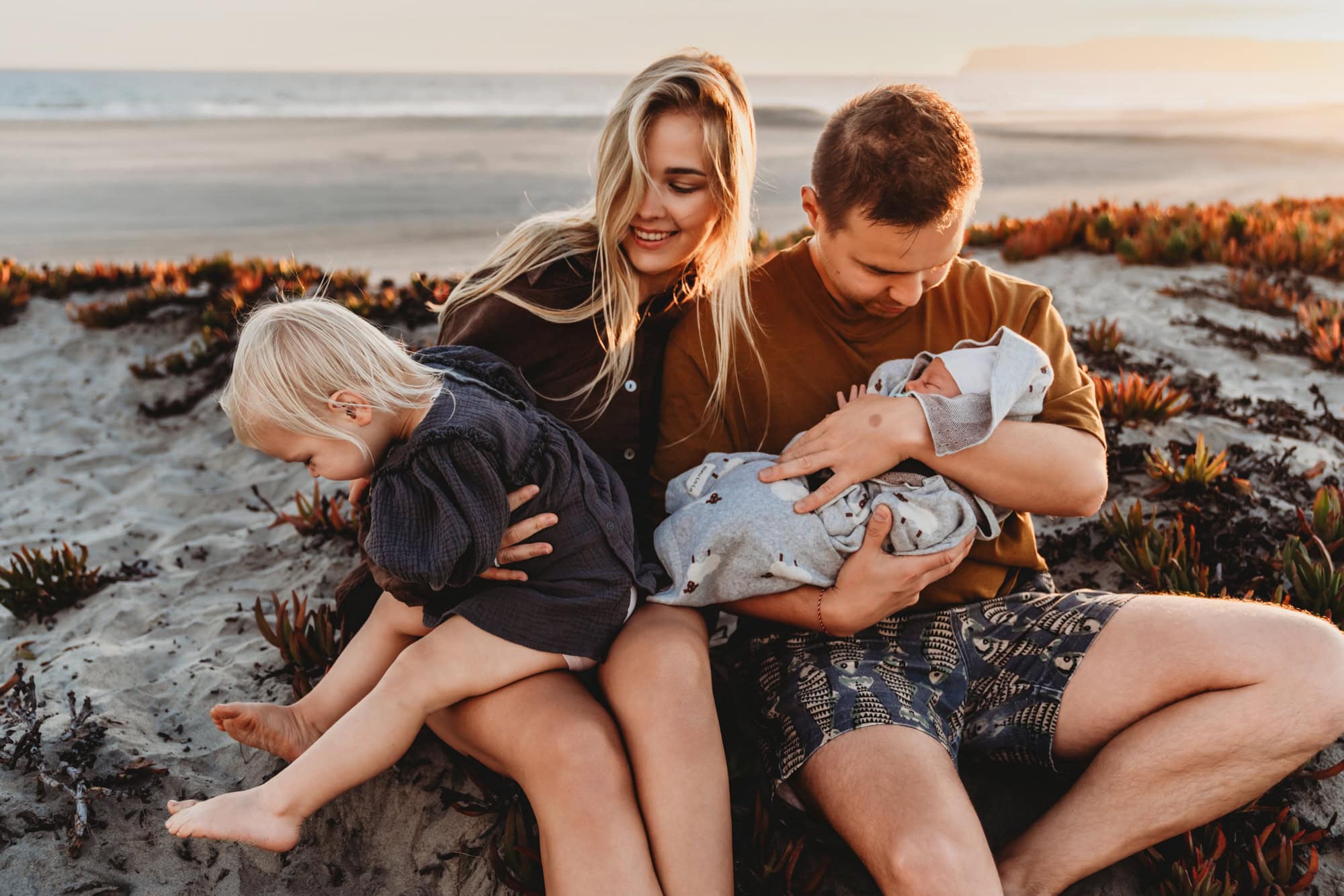 Parents sitting on the sand at Coronado Beach, each cradling the newborn while big sister leans in during an outdoor newborn photoshoot in San Diego