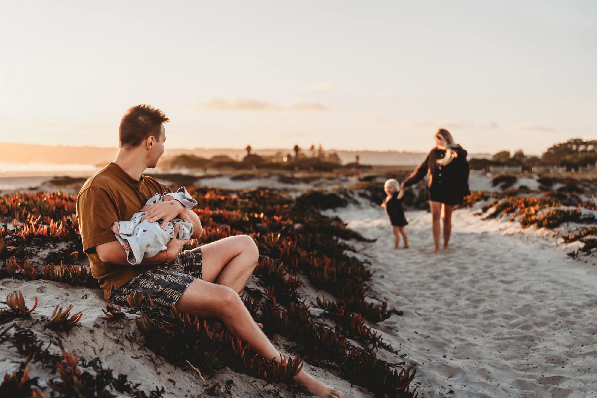Dad with baby, mom with toddler in distance Father sitting on the sand holding his newborn while mom walks with their toddler along Coronado Beach during a relaxed San Diego outdoor newborn session.