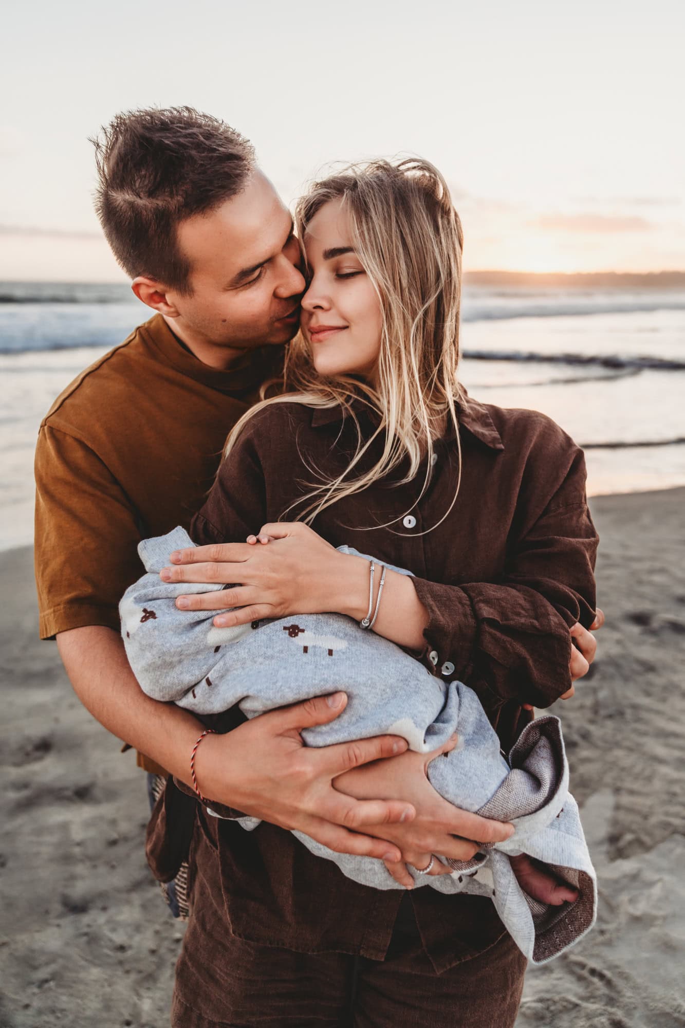 Parents embracing with newborn by the water Parents wrapped in an embrace, holding their newborn near the San Diego shoreline during a golden-hour outdoor newborn session in San Diego.
