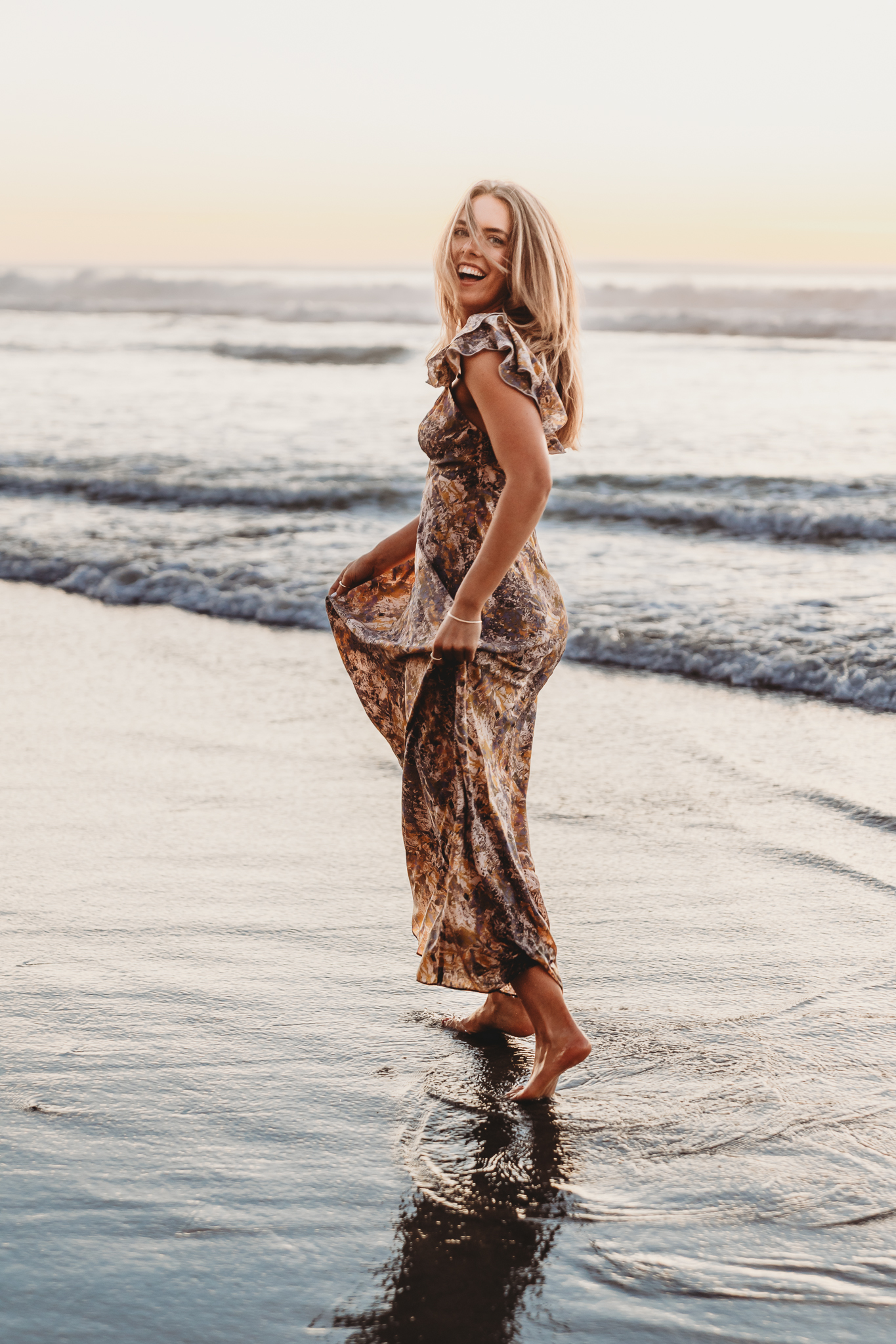 A senior having her senior portraits taken plays in the surf at Torrey Pines State Beach in San Diego with soft ocean light and shoreline reflections.