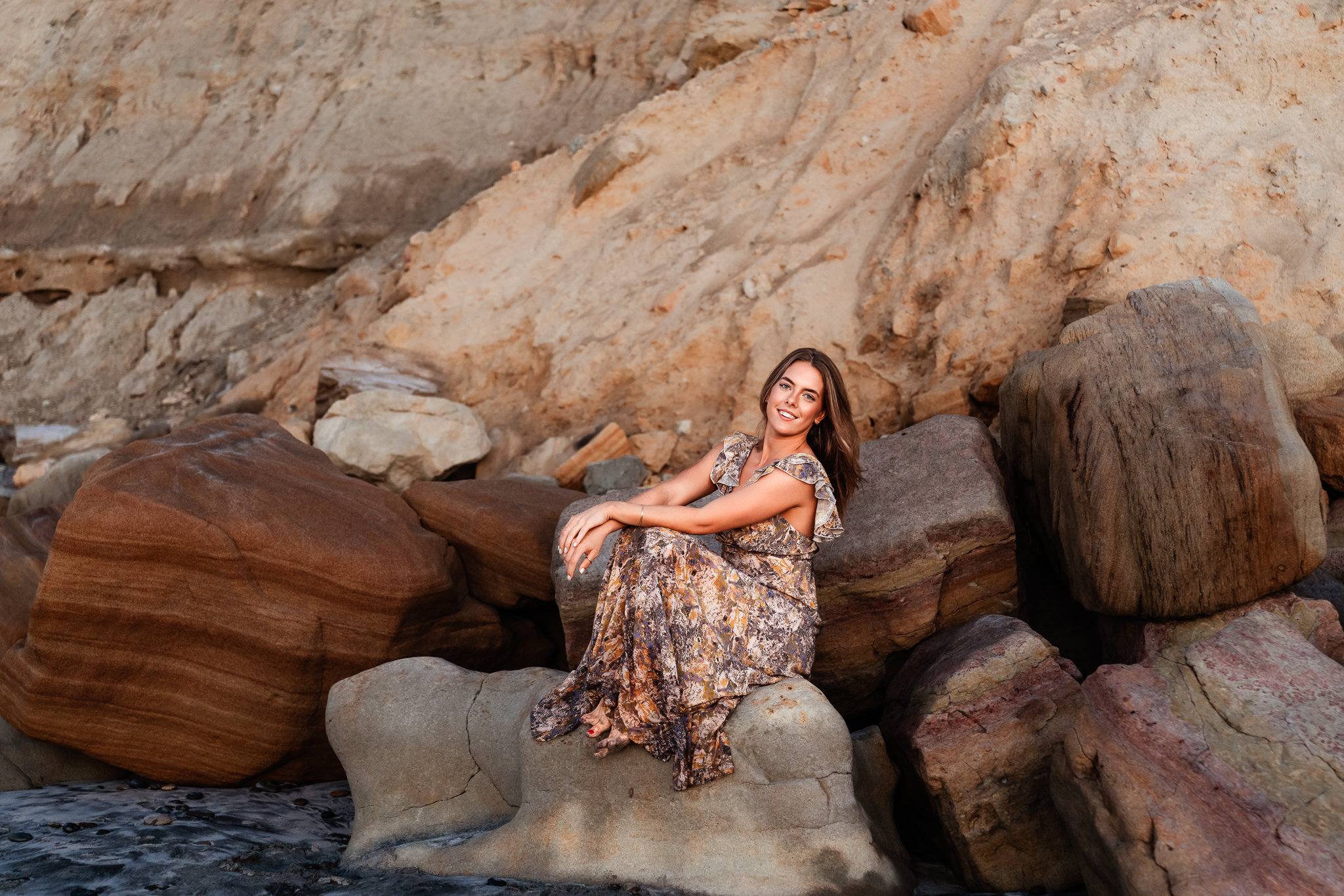 A senior girl sits on rocks, looking at the camera and smiling, on Torrey Pines beach during her senior photo session in San Diego.