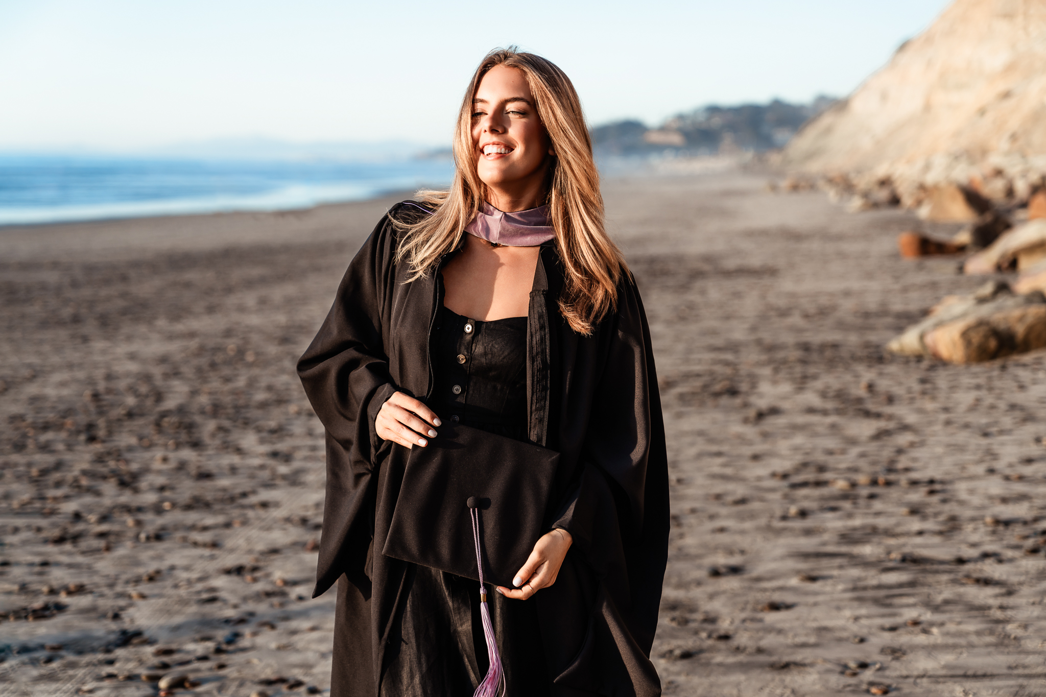 High school senior holds her cap while wearing her gown during her senior portraits session at Torrey Pines State Beach in San Diego.