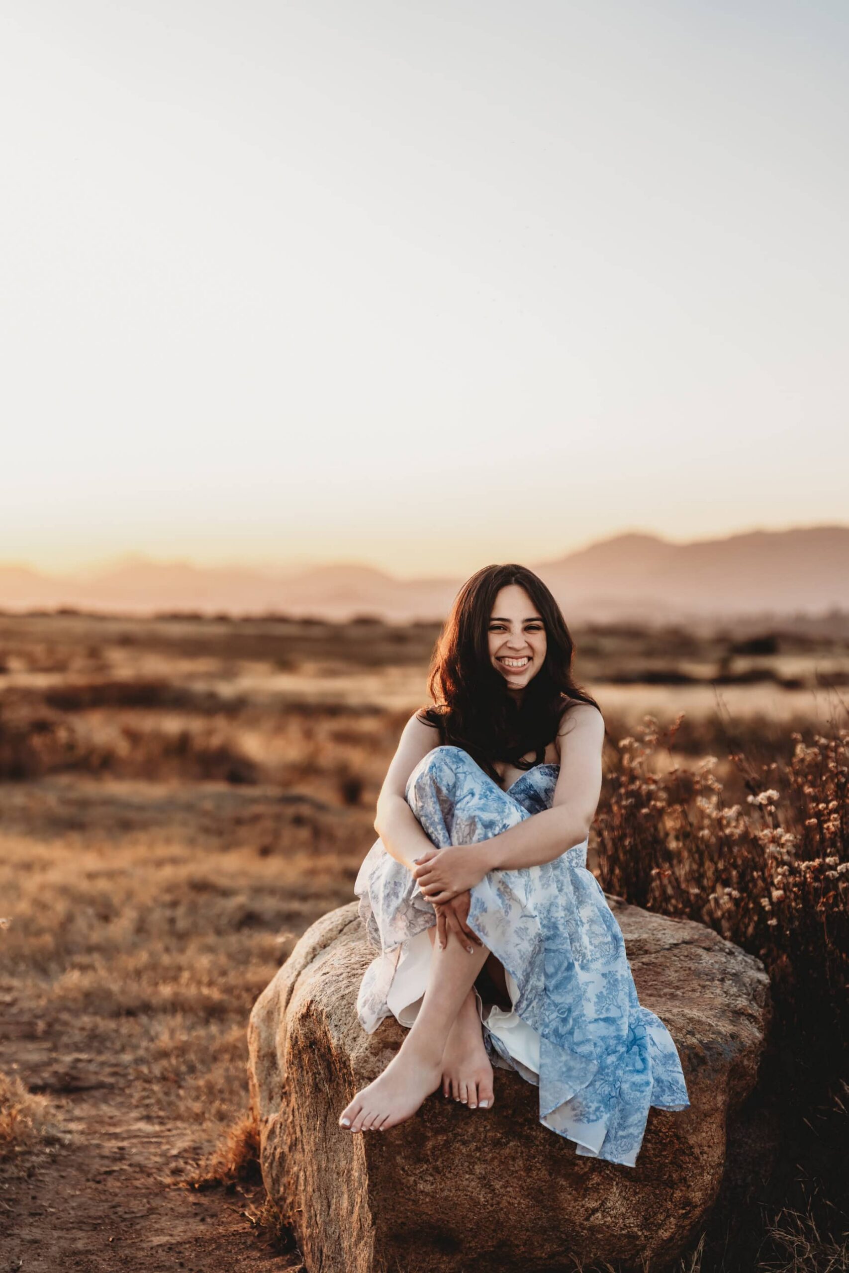 High school senior in a blue and white dress sitting on a rock in a field with mountains in the background at golden hour, San Diego senior photos.