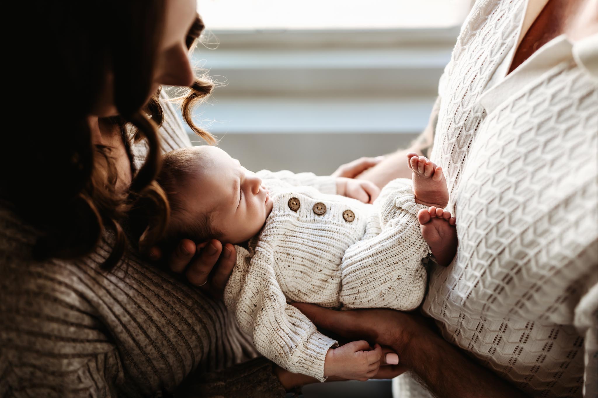 Close-up lifestyle newborn photo of a sleeping baby in a knit outfit being cradled by parents in warm window light during an in-home session in San Diego