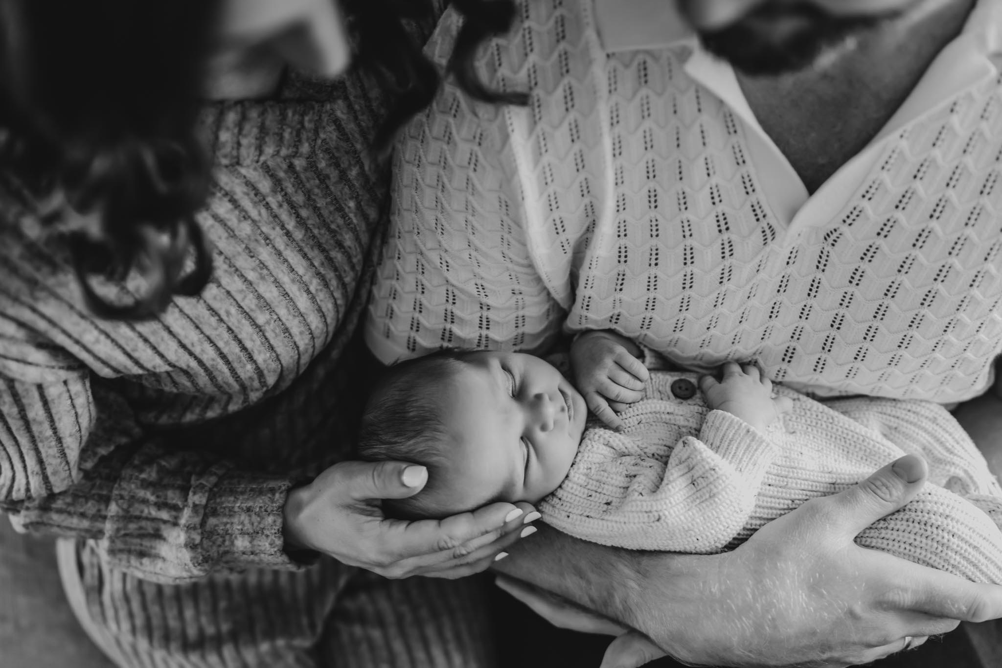 Overhead black and white photo of parents' hands gently cradling their newborn baby during a lifestyle newborn session in San Diego