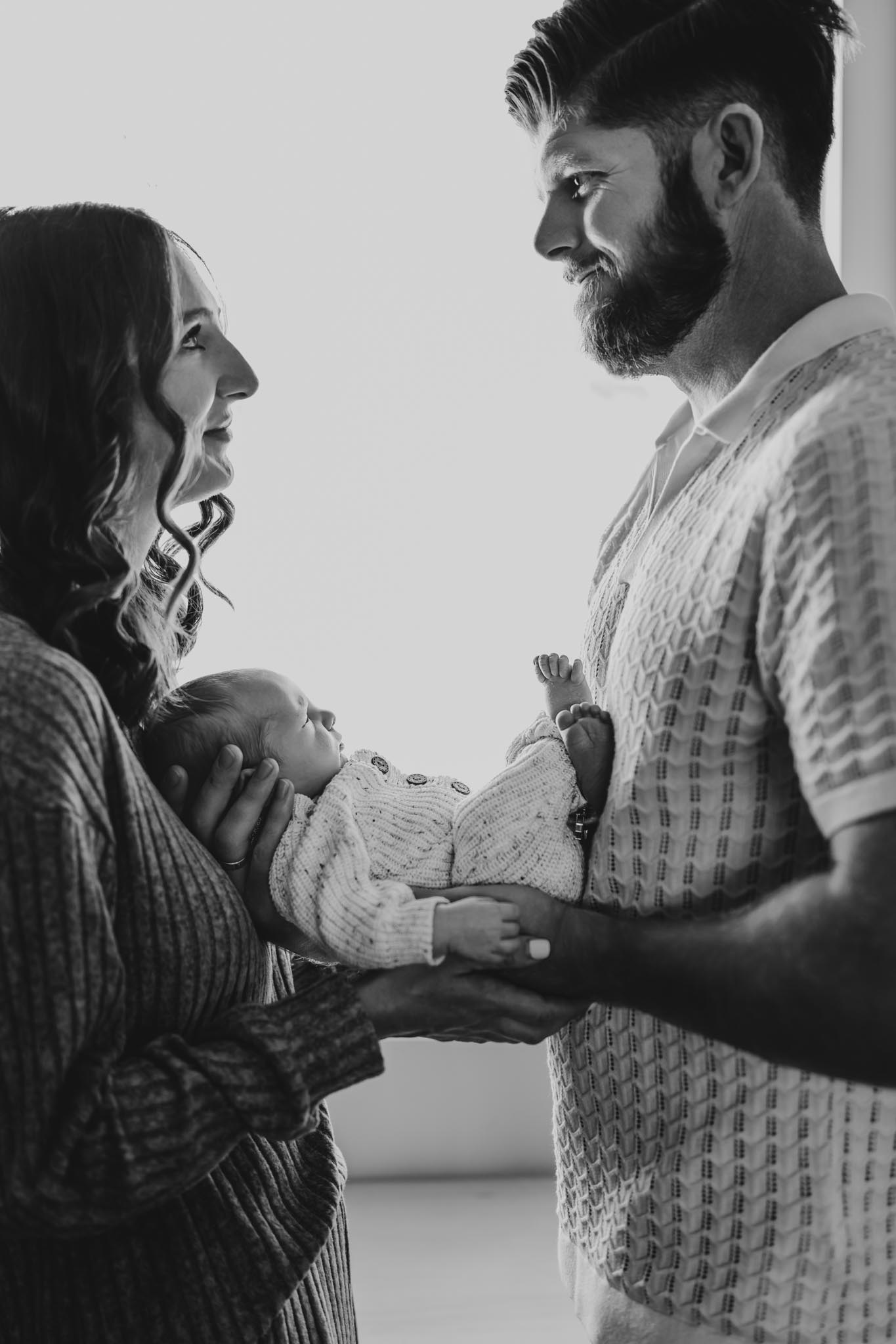 Black and white lifestyle newborn photo of mom and dad facing each other and smiling, holding their newborn baby in soft window light, San Diego