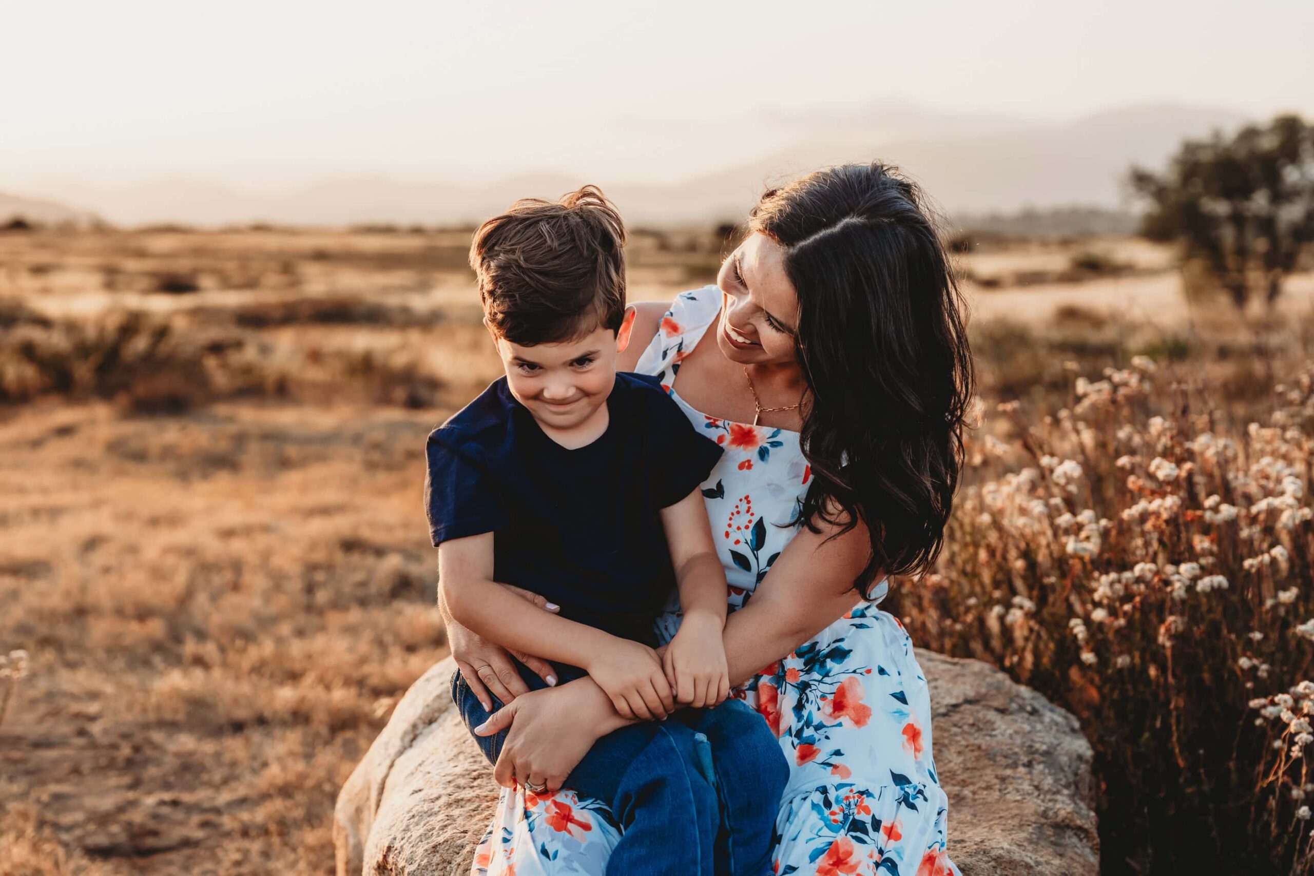 Mom and son laughing together while sitting on a rock in a golden San Diego field