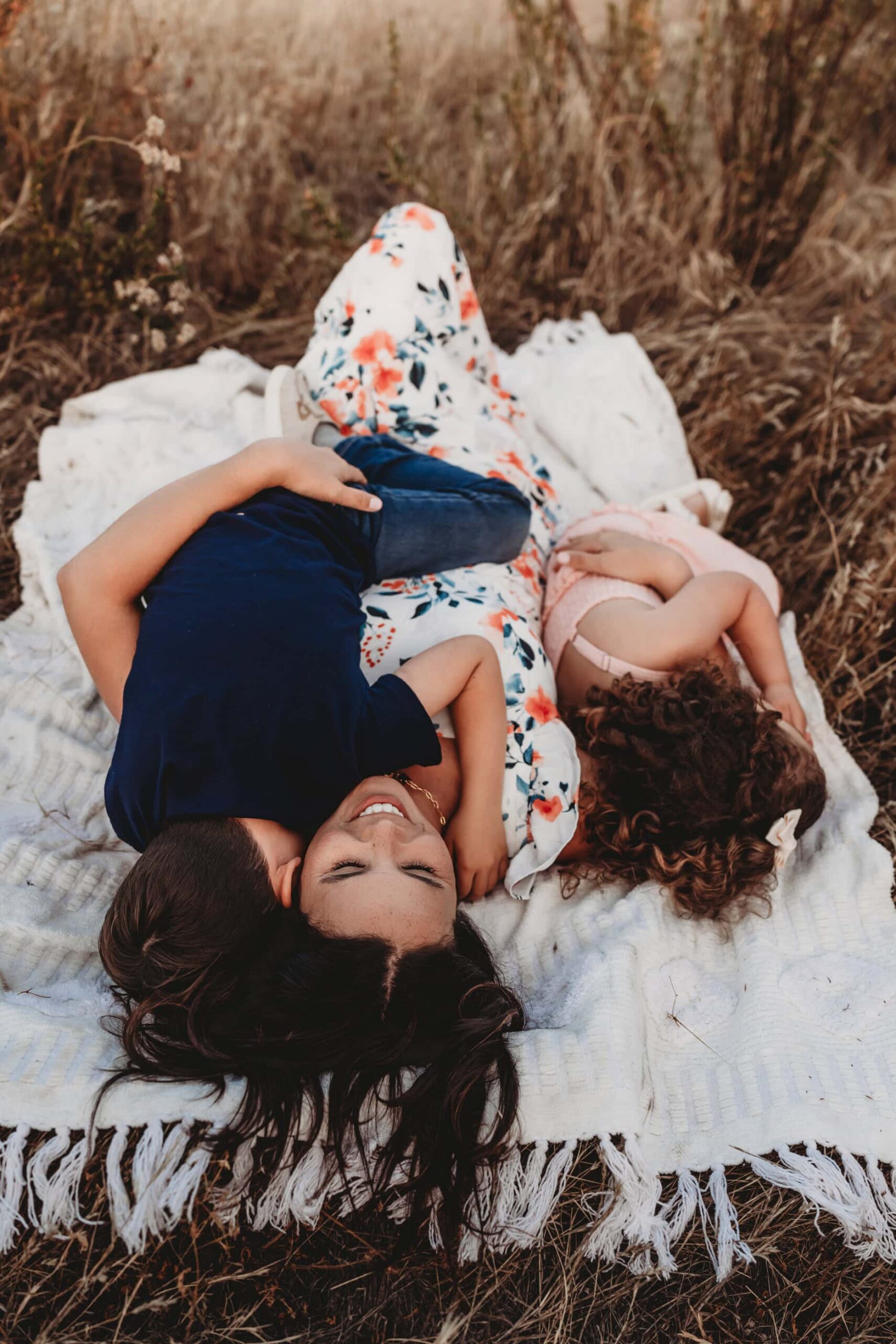 Mom lying on a blanket in a golden field, laughing with her two children during a family photo session